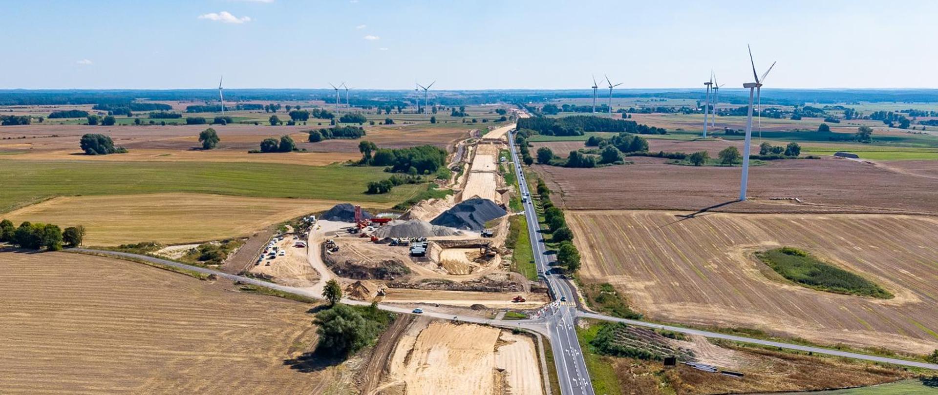 Widok z lotu ptaka na budowę drogi ekspresowej przebiegającą przez rozległy krajobraz rolniczy. W centrum zdjęcia widoczny jest przygotowany korytarz przyszłej trasy z nasypami ziemnymi, sprzętem budowlanym i składowiskami kruszywa. Obok placu budowy biegnie istniejąca droga. Po obu stronach rozciągają się pola uprawne w różnych odcieniach zieleni i brązu oraz pojedyncze zadrzewienia. W tle znajdują się liczne elektrownie wiatrowe rozmieszczone na otwartej równinie. Zdjęcie wykonano w słoneczny dzień przy dobrej widoczności.