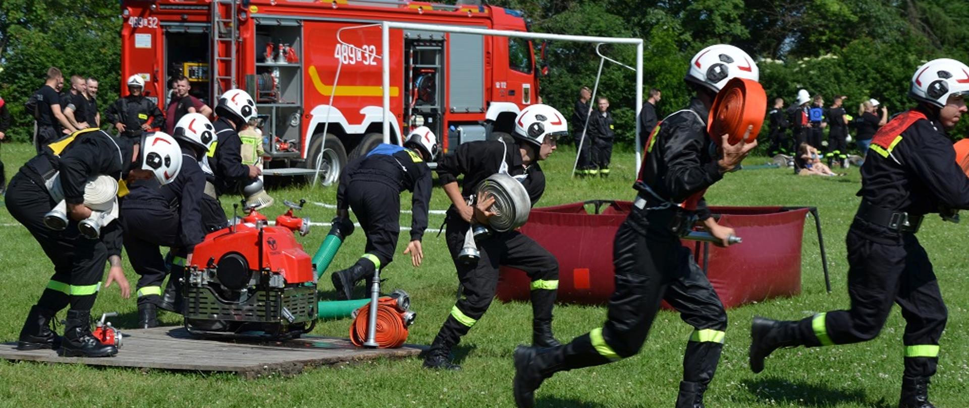 Strażacy podczas wykonywania ćwiczenia bojowego. W tle stoi samochód pożarniczy