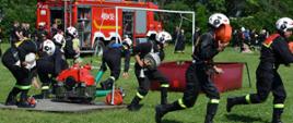 Strażacy podczas wykonywania ćwiczenia bojowego. W tle stoi samochód pożarniczy