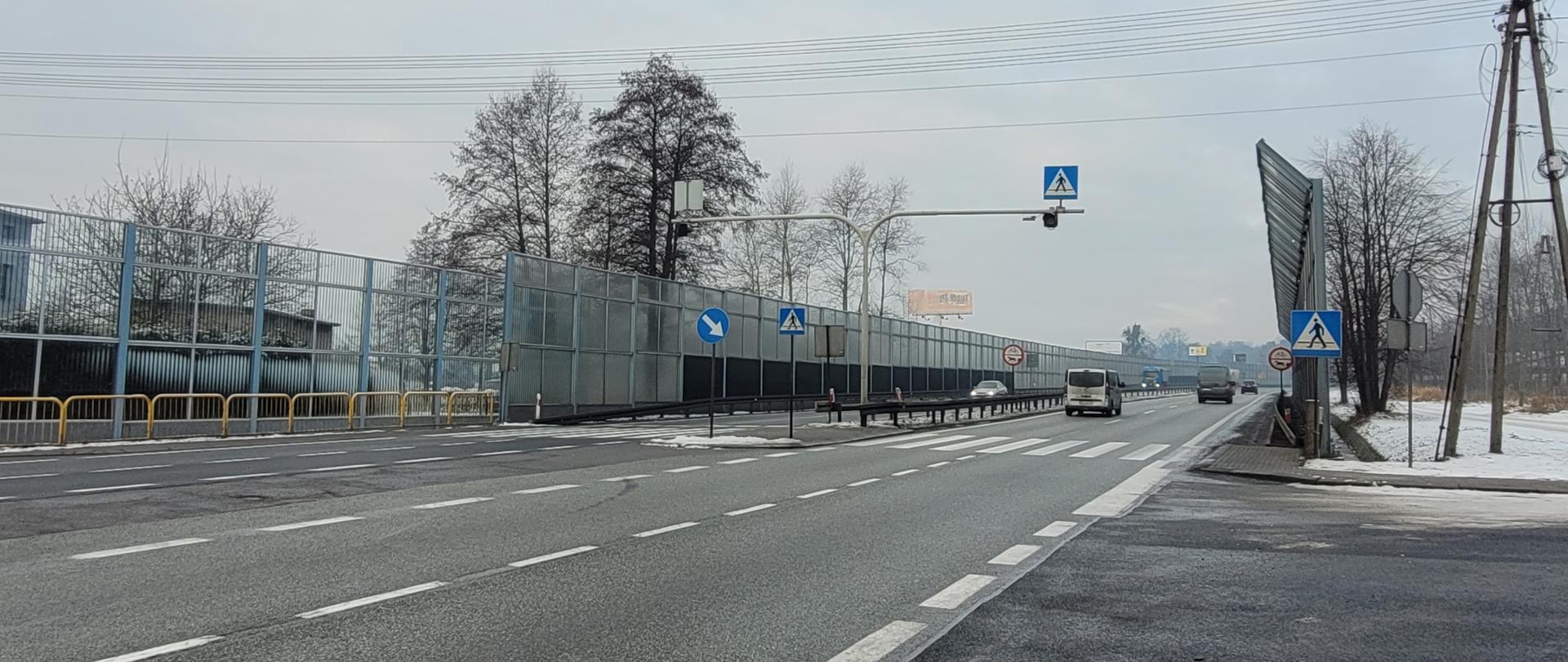 Fotografia z poziomu terenu przejścia przez DK1 w rejonie skrzyżowania z ul. Podleśną w Piasku w aurze zimowej. Na dwujezdniowej drodze z dwoma pasami w każdym z kierunków poruszają się pojazdy. Jezdnie rozdzielone pasem zieleni wyposażonym w bariery ochronne, przejście dla pieszych we wsypę separacyjną. 