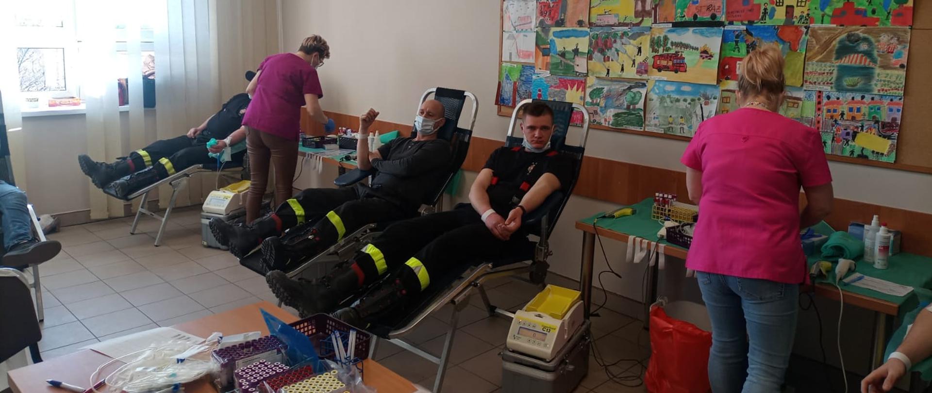 Zdjęcie przedstawia trzech strażaków oddających honorowo krew. Tyłem do zdjęcia dwie pielęgniarki obsługujące oddających. Za nimi tablica z kolorowymi rysunkami wykonanymi przez dzieci.