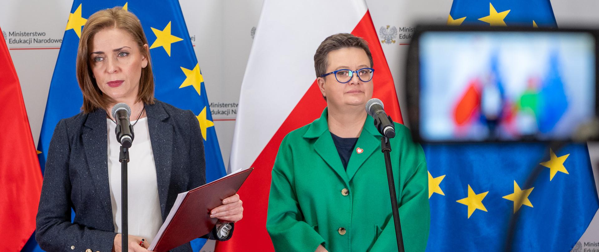 Wiceminister Katarzyna Lubnauer i Joanna Mucha stoją przed ustawionymi na statywach mikrofonami. W tle flagi Polski i UE