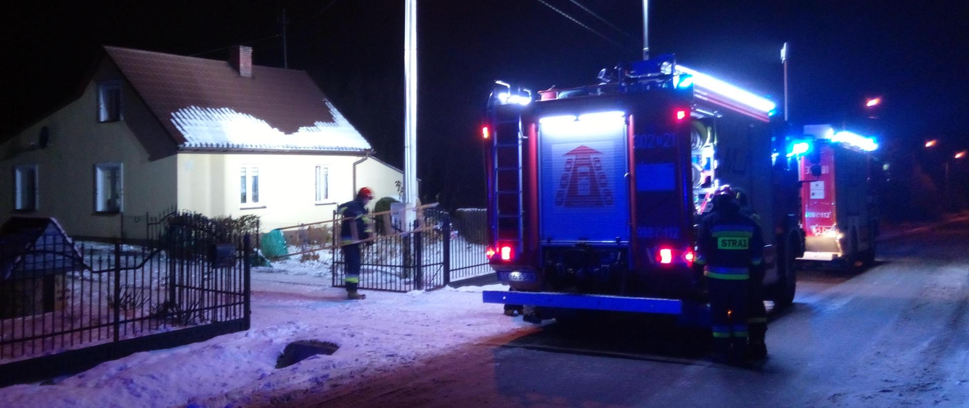 Fotografia przedstawia samochód pożarniczy na pierwszym planie, na drugim planie znajduje się budynek mieszkalny w którym doszło do pożaru. Nie widać dymu ani ognia. Strażak przy bramie wjazdowej trzyma drabinę.