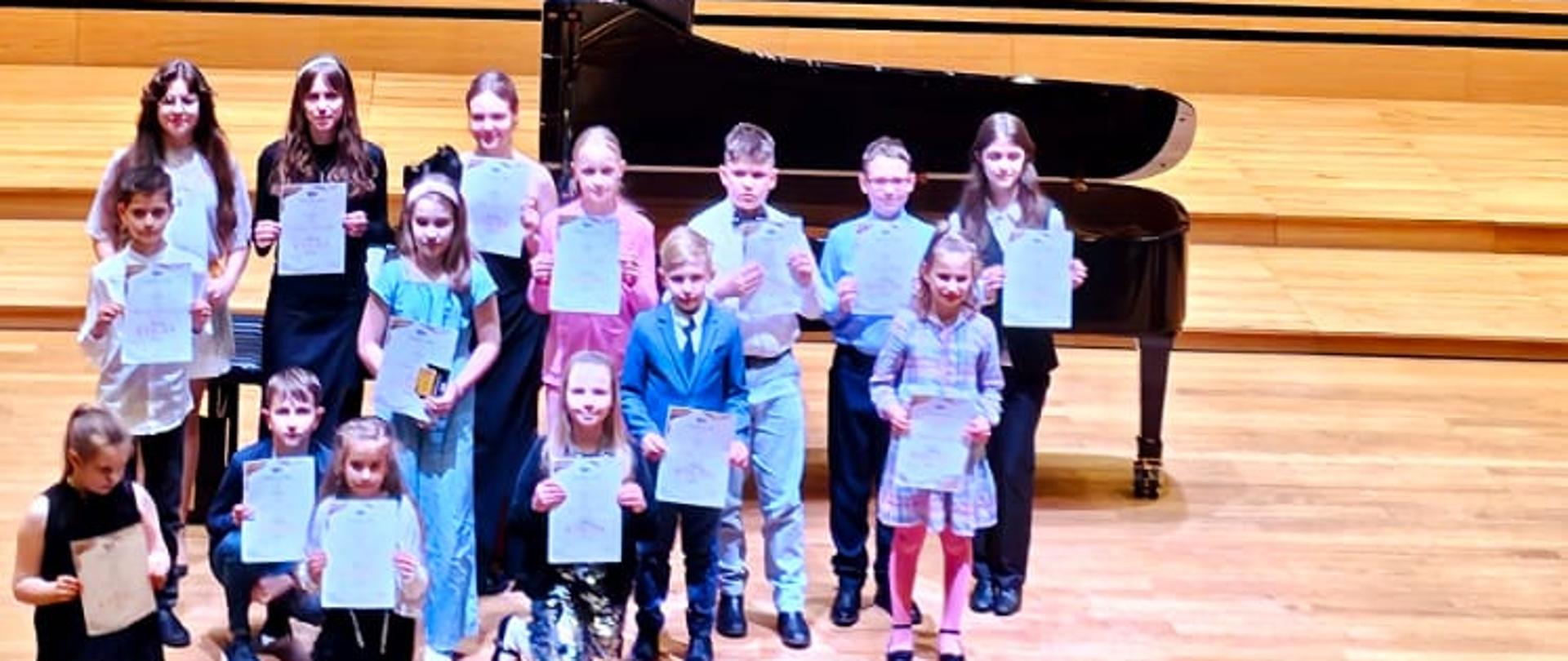 Dzieci i młodzież stoją w dwóch rzędach na scenie trzymając w rękach dyplomy.