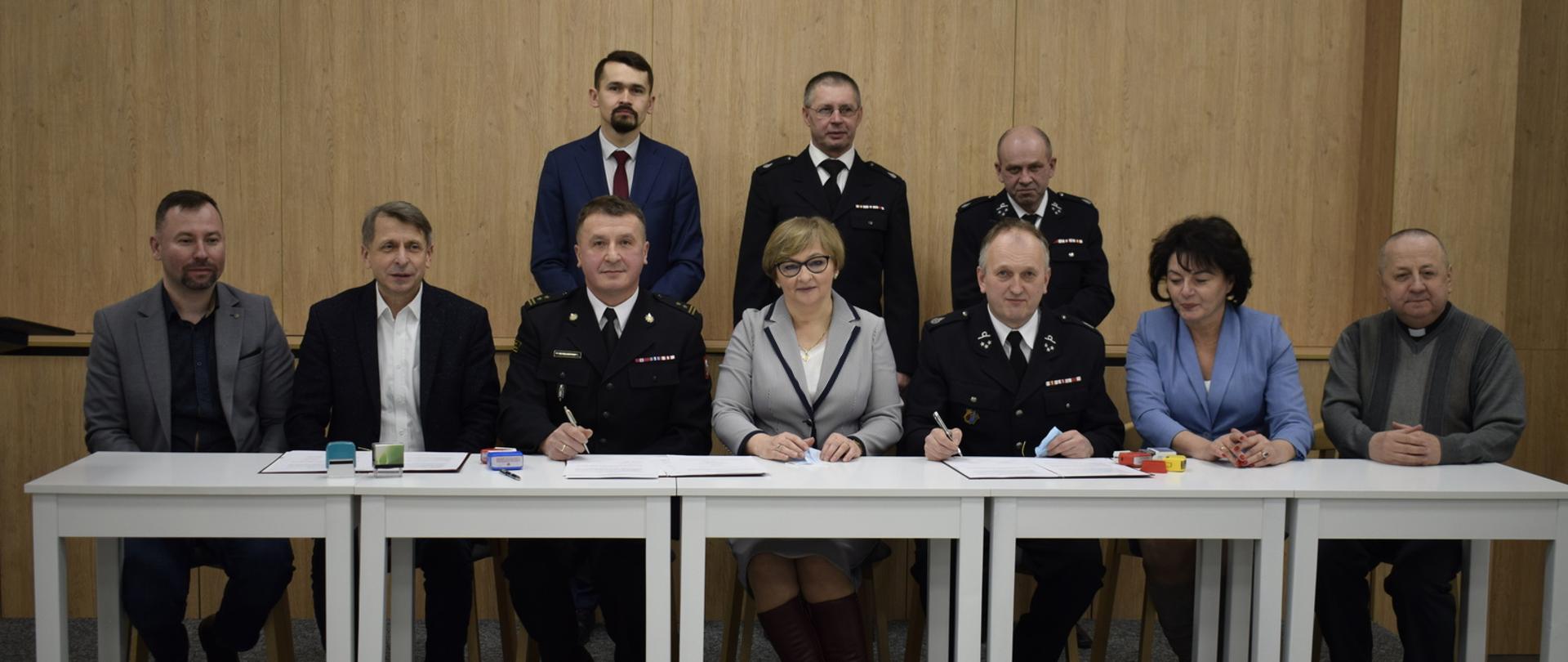 Na zdjęciu widoczne 10 osób cztery osoby w mundurach strażackich pozostałe to osoby cywilne w tym dwie kobiety. Cześć osób siedzi przy stoliku prezydialnym i podpisują porozumienie weryfikujące włączenie jednostki OSP w Rożnowicach do KSRG