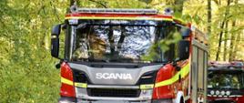 Czerwono-srebrny wóz strażacki marki Scania z napisem „OSP TRĘBACZÓW” jedzie leśną, asfaltową drogą. Pojazd ma włączone światła. Za nim podąża kolejny czerwony wóz strażacki. W tle widać zielone i żółknące drzewa.