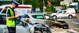 Na zdjęciu widoczny jest funkcjonariusz Policji. Na drugim planie widoczne są dwa uszkodzone pojazdy w wyniku zdarzenia drogowego. Na zdjęciu w oddali widoczna jest również karetka pogotowia oraz samochód straży pożarnej.