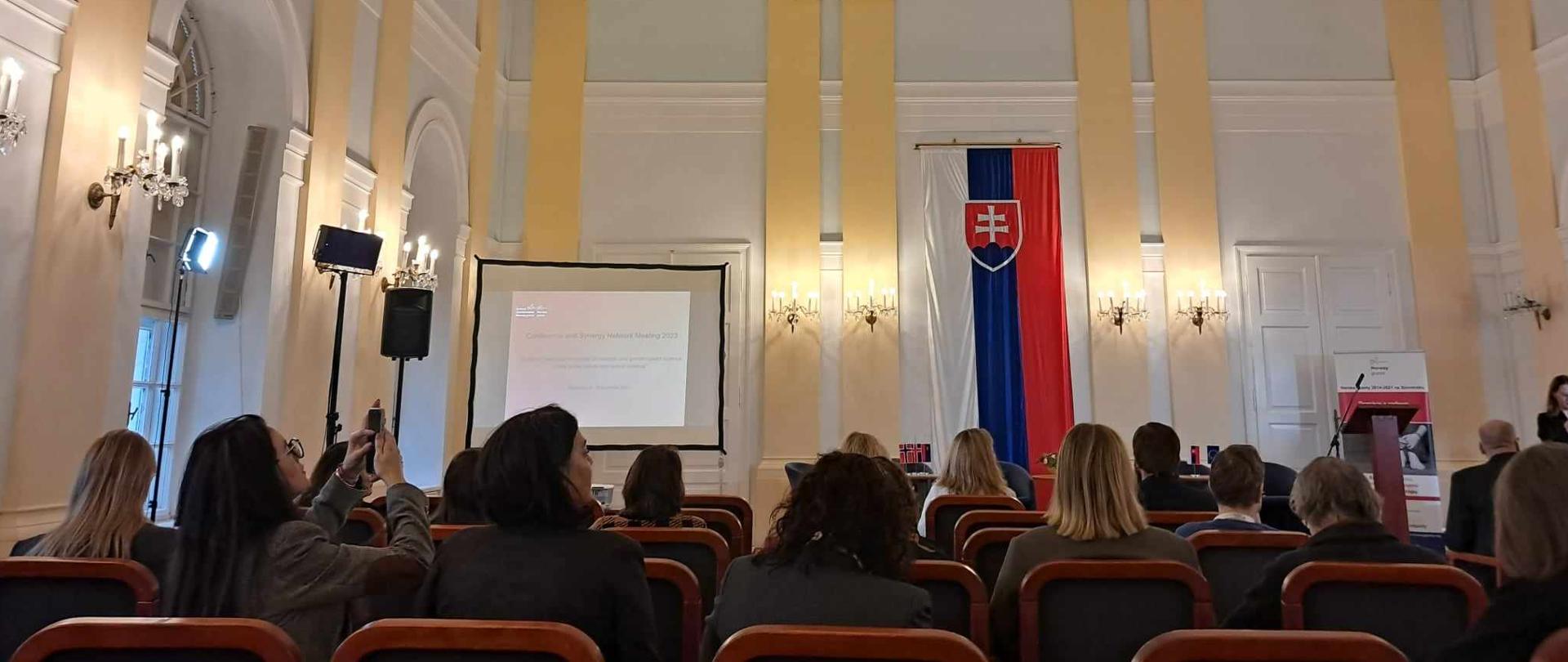 Na pierwszym planie widać siedzące osoby uczestniczące w konferencji. Na drugim planie widać flagę oraz mównicę. 