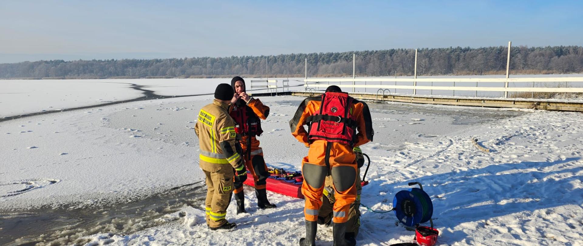 Strażacy przygotowują sprzęt potrzebny do przeprowadzenia ćwiczeń na lodzie. Ubierani są w kamizelki asekuracyjne, kaski oraz w ubrania do pracy w wodzie
