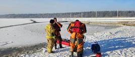 Strażacy przygotowują sprzęt potrzebny do przeprowadzenia ćwiczeń na lodzie. Ubierani są w kamizelki asekuracyjne, kaski oraz w ubrania do pracy w wodzie

