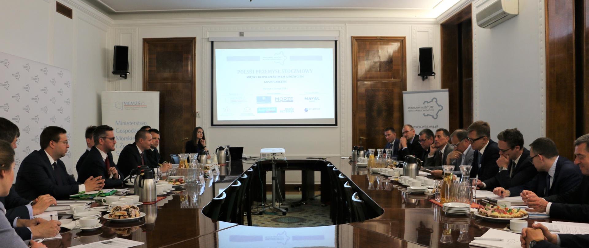 Uczestnicy warsztatów siedzą przy okrągłym stole. W tle, na rzutniku widoczna jest prezentacja dotycząca omawianej tematyki.