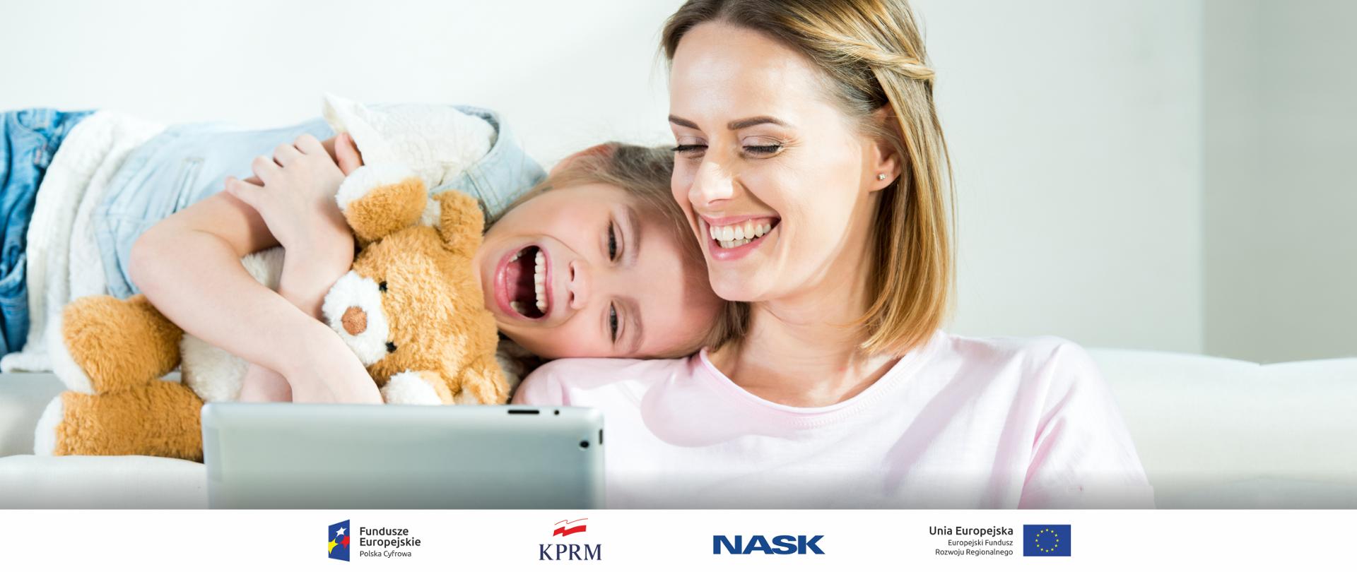 Zdjęcie uśmiechniętych mamy i córeczki. Dziewczynka opiera głowę na ramieniu kobiety. Kobieta korzysta z laptopa, dziewczynka tuli misia.