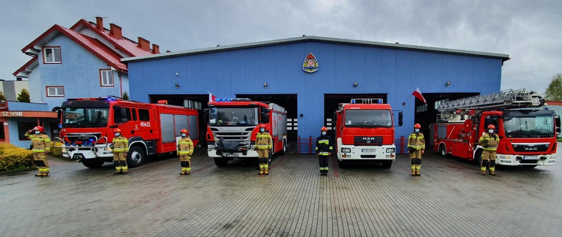 Zdjęcie przedstawia 4 samochody specjalne Państwowej Straży Pożarnej dla tle garażu jednostki ratowniczo-gaśniczej w Jędrzejowie. Przed pojazdami znajdują się strażacy w umundurowaniu specjalnym z założonymi czerwonymi hełmami na głowie oraz maseczkami ochronnymi zakrywającymi usta i nos. Strażacy oddają hołd tragicznie zmarłemu policjantowi.