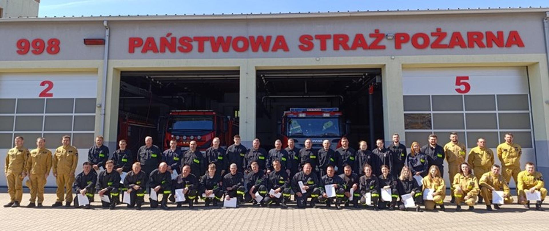 Zdjęcie przedstawia dużą grupę strażaków, stojących i klęczących w dwóch rzędach przed budynkiem Państwowej Straży Pożarnej. Na froncie budynku widnieje napis "PAŃSTWOWA STRAŻ POŻARNA" oraz numery bram garażowych "2" i "5". W tle, przez otwarte bramy garażowe, widać zaparkowane czerwone wozy strażackie. W pierwszym rzędzie, na pierwszym planie, strażacy klęczą i trzymają w rękach białe dokumenty, prawdopodobnie dyplomy lub zaświadczenia. Ubrani są w ciemne (czarne) mundury. W drugim rzędzie stoją strażacy w jasnobeżowych (bojowych) mundurach. Scena sugeruje uroczystość ukończenia szkolenia.