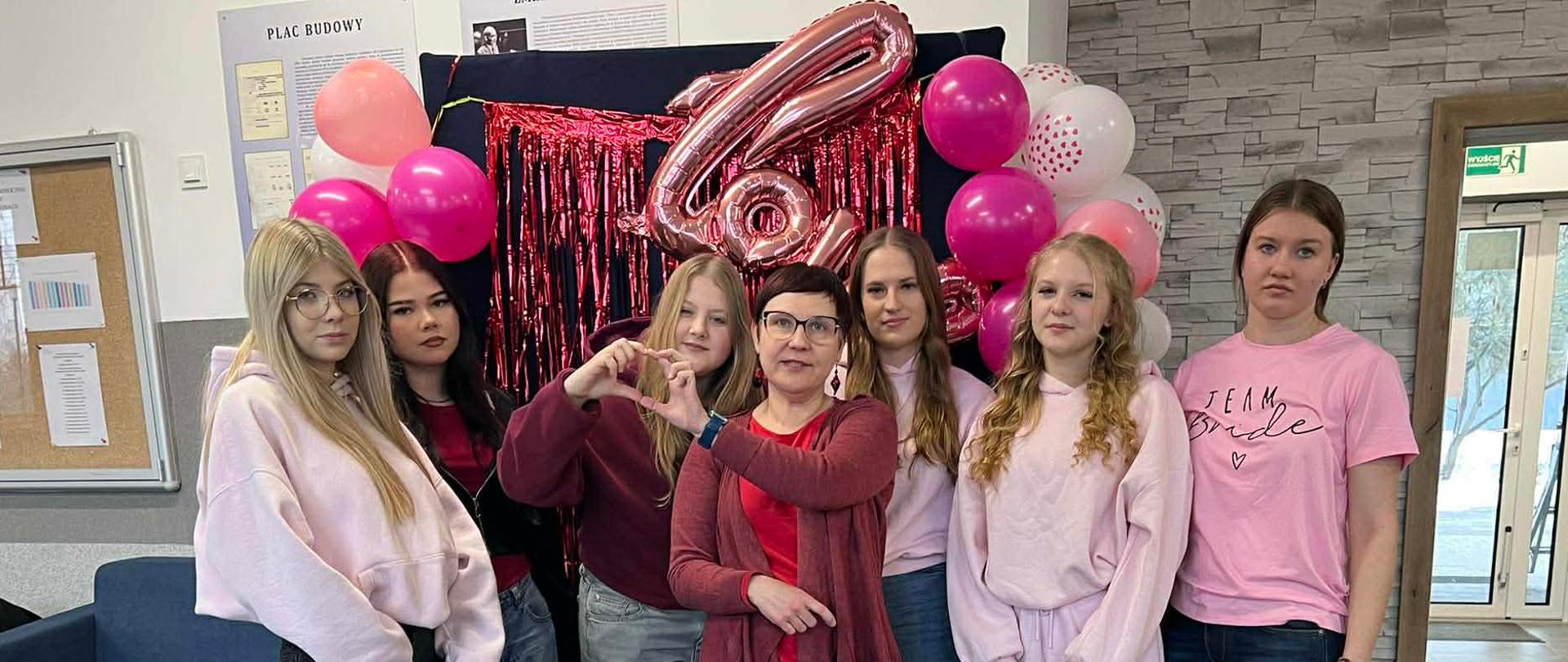 Grupa dziewcząt i opiekunka pozują do pamiątkowego zdjęcia na tle różowych balonów i błyszczących dekoracji. Osoby w centrum i na pierwszym planie układają dłonie w kształt serca.