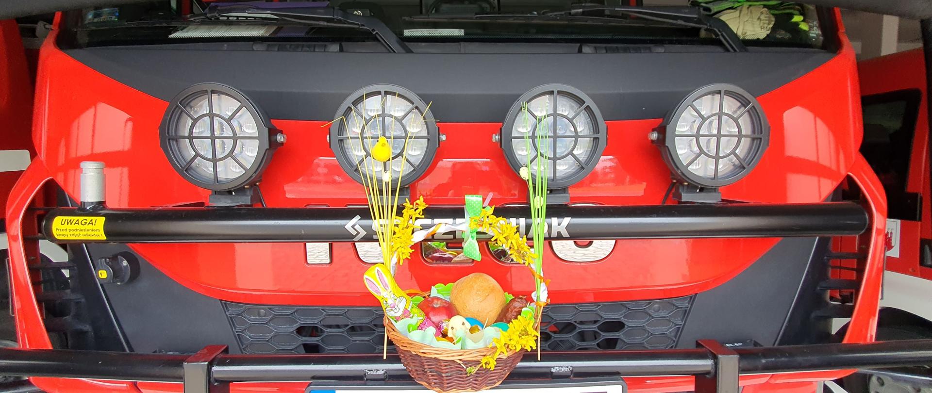 Zdjęcie przedstawia przód czerwonego pojazdu strażackiego z widocznymi 4 reflektorami. przed nim stoi wielkanocny koszyczek, w którym znajduje się zając baranek, owoce, pieczywo i pisanki.
