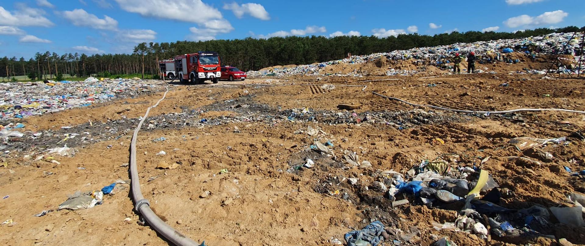 Składowisko odpadów, widoczne składowane śmieci, w oddali wóz strażacki. Na pierwszym planie wąż strażacki. 
