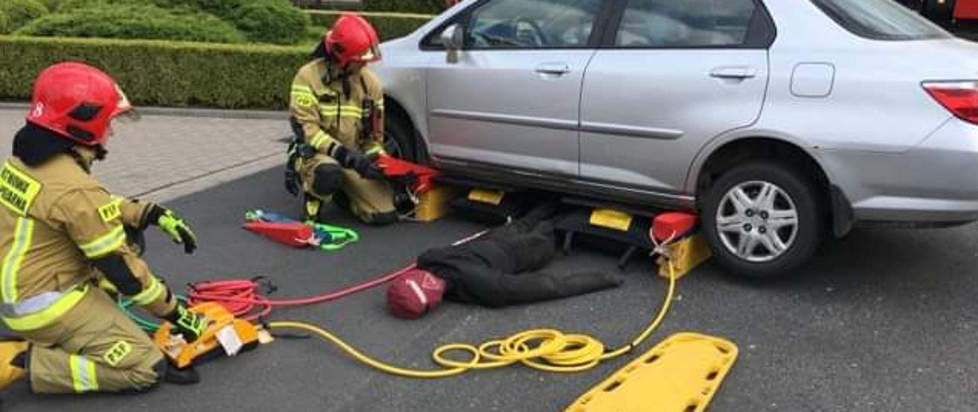 Dwóch strażaków podczas doskonalenia zawodowego z ratownictwa technicznego. Za pomocą poduszek pneumatycznych podnoszą samochód pod, którym umieszczony jest fantom do pozoracji