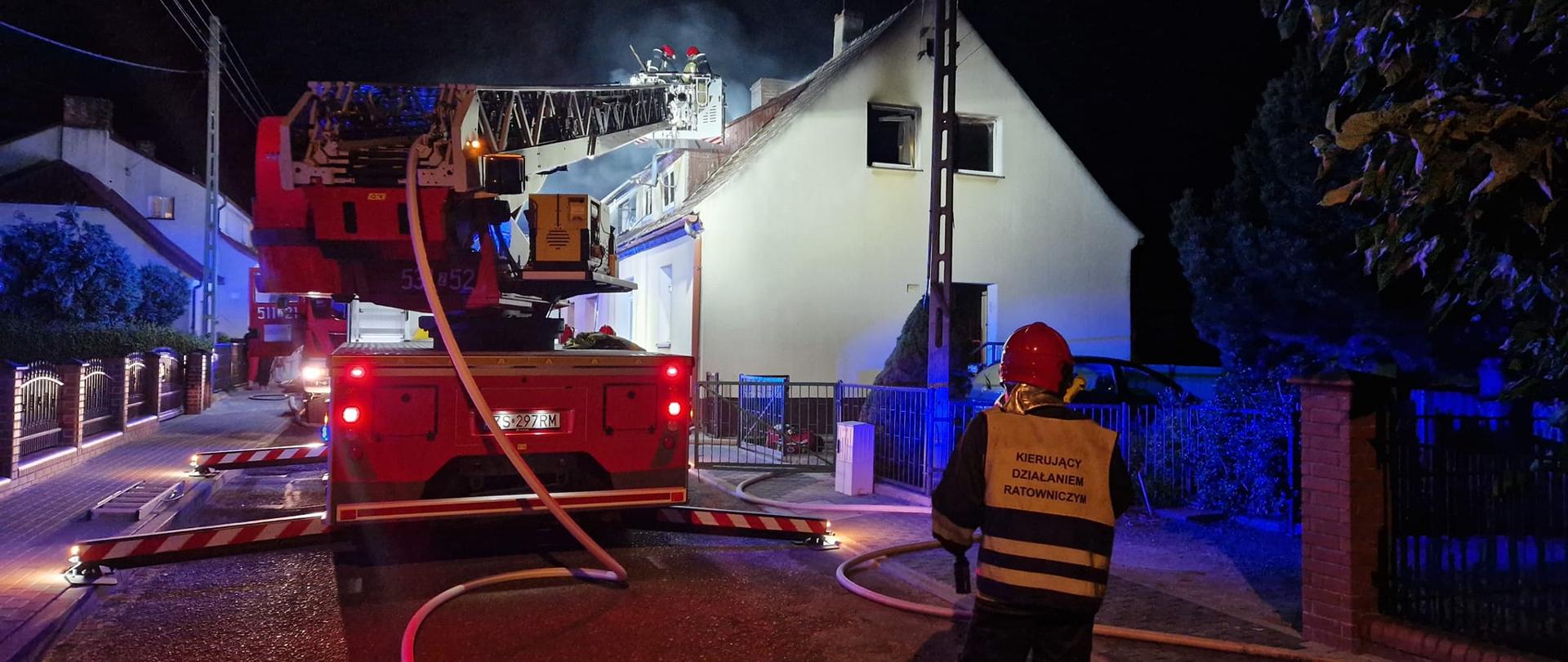 Na zdjęciu stojący tyłem strażak kierujący działaniem ratowniczym oraz drabina pożarnicza, w koszu drabiny dwóch strażaków gaszących pożar poddasza budynku mieszkalnego.