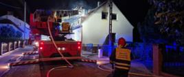 Na zdjęciu stojący tyłem strażak kierujący działaniem ratowniczym oraz drabina pożarnicza, w koszu drabiny dwóch strażaków gaszących pożar poddasza budynku mieszkalnego.