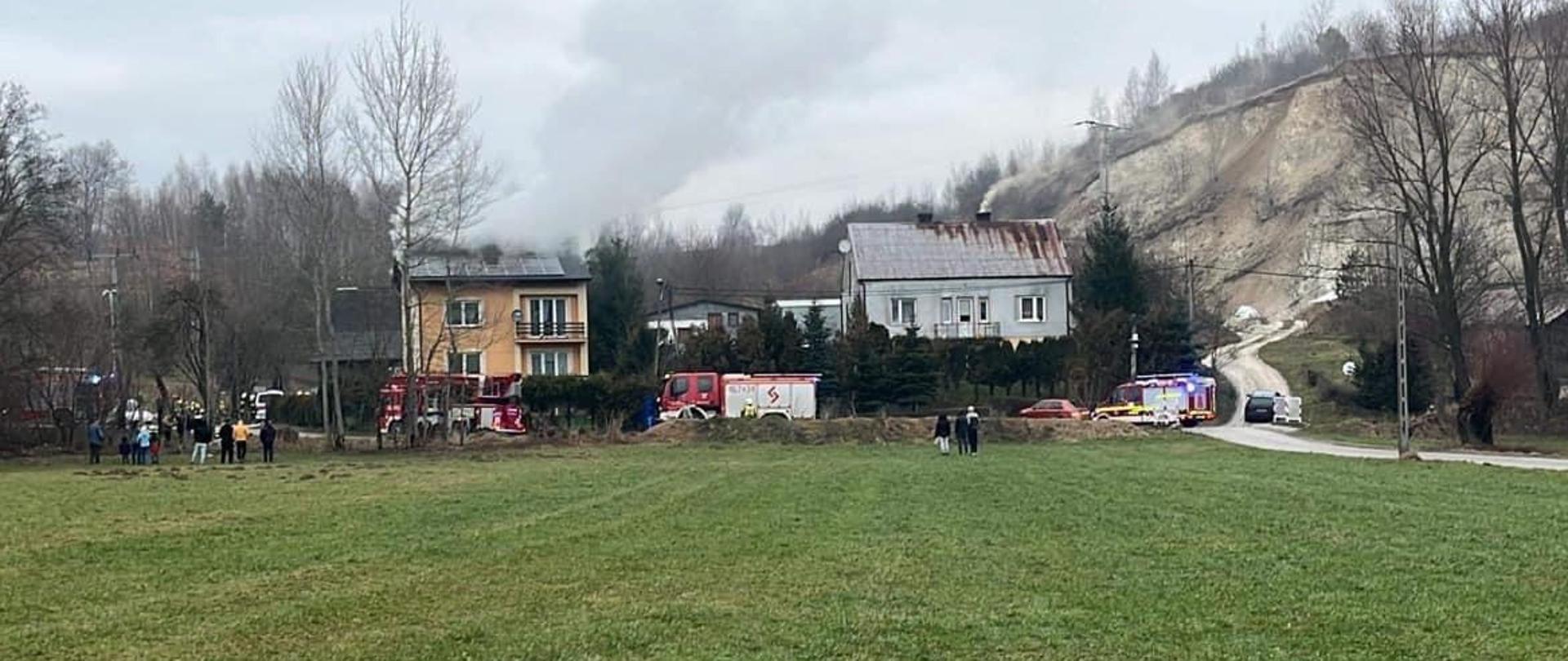 Na zdjęciu widoczny dom mieszkalny z którego wydobywa się dym obok zaparkowane samochody pożarnicze