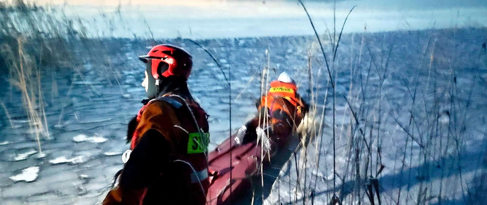 Zdjęcie przedstawia strażaków, którzy za pomocą sań lodowych pokonują zamarzniętą taflę jeziora. Na saniach leży strażak w stroju do pracy w wodzie, drugi go asekuruję. W tle widać zamarznięte jezioro, trzciny.