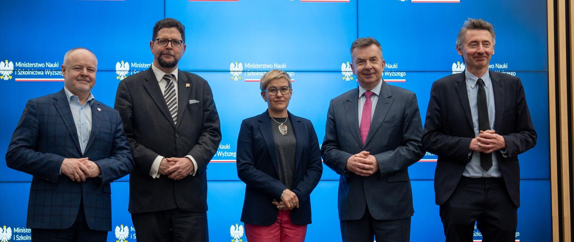 Minister Wieczorek i wiceministrowie stoją na sali, pod wielkim wbudowanym w ścianę ekranem.