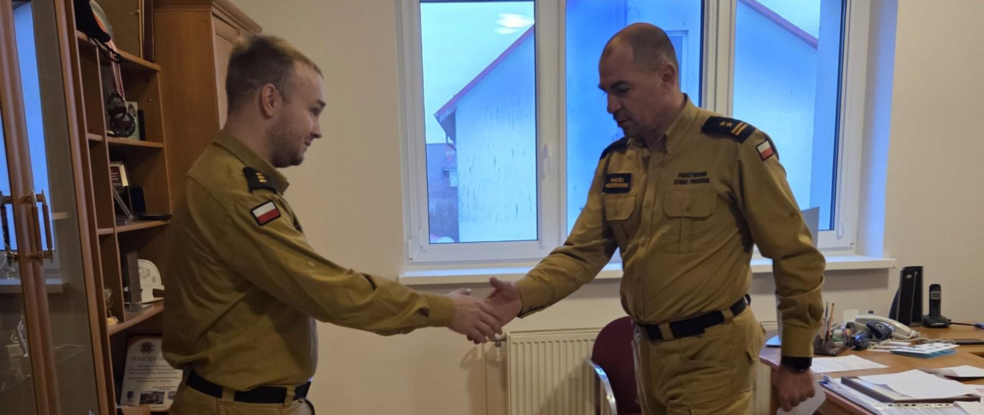 W biurze dwóch strażaków w służbowych mundurach stoi naprzeciwko siebie. Jeden z nich trzyma w ręku dokument i odczytuje go, podczas gdy drugi stoi w postawie wyprostowanej, słuchając. W pomieszczeniu widać biurko z dokumentami, regał oraz okno z widokiem na budynki na zewnątrz