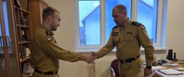 Ci sami dwaj strażacy w mundurach podają sobie ręce w geście gratulacji lub przekazania wyróżnienia. Jeden z nich nadal trzyma w drugiej ręce dokument. Scena odbywa się w tym samym biurze, przy biurku i regale stojącym obok.