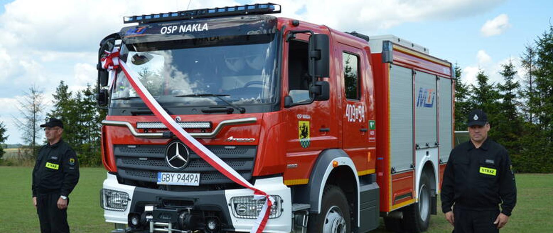 Zdjęcie przedstawia średni samochód ratowniczo-gaśniczy marki Mercedes Benz koloru czerwonego z przewieszoną biało-czerwoną wstęgą przez przednią szybę oraz ustawionych po obu stronach pojazdu strażaków w czarnym umundurowaniu koszarowym