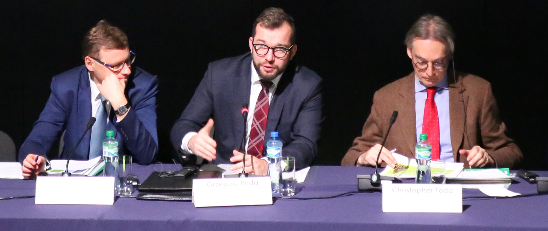 Przy stole przykrytym fioletową tkaniną siedzi trzech mężczyzn. Pośrodku wiceminister Grzegorz Puda.