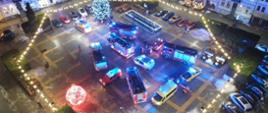 Zdjęcie wykonane z góry z drona. Przedstawia pojazdy ratownicze ułożone w kształcie choinki. Zdjęcie oświetlone lampami miejskimi i błyskami pojazdów.