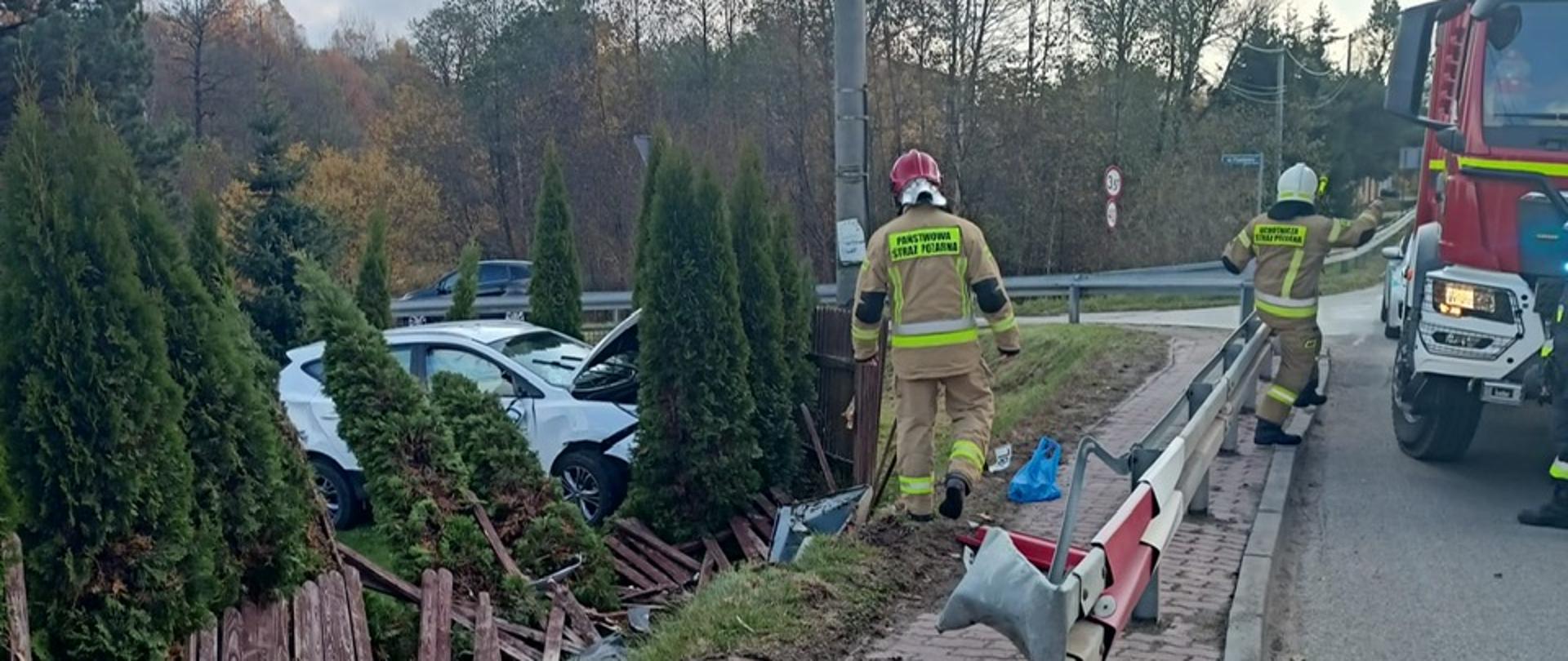 Zdjęcie przedstawia biały samochód osobowy stojący wśród ozdobnych krzewów na terenie prywatnej posesji. Widać zniszczone ogrodzenie posesji oraz barierki energochłonne. Chodnikiem idzie strażak, drugi wsiada do samochodu pożarniczego.