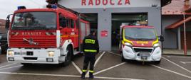 Strażak OSP stoi przed dwoma samochodami pożarniczymi, które są zaparkowane przed remizą OSP.