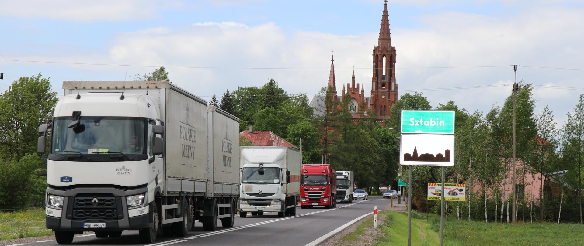 Ciąg czterech samochodów ciężarowych na jednojezdniowej drodze. Po prawej stronie znak z nazwą miejscowości Sztabin i oznaczeniem terenu zabudowanego, w tle wieża kościelna