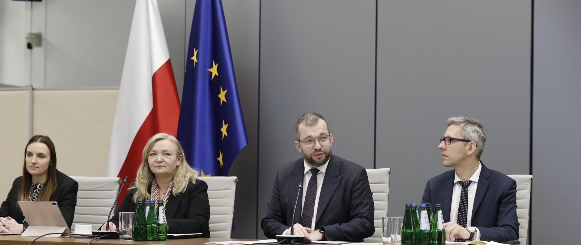 Cztery osoby siedzą na krzesłach obok siebie. Jako drugi z prawej strony siedzi minister Grzegorz Puda.