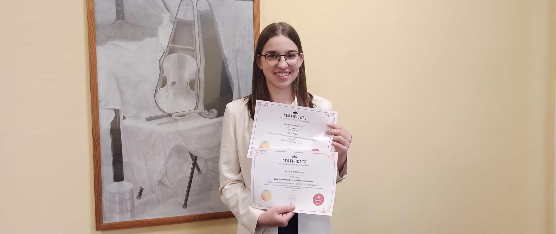 Zdjęcie kolorowe. Ciemnowłosa dziewczyna w jasnym żakiecie trzyma w rękach konkursowe dyplomy. W tle czarno - biały obraz - martwa natura ze skrzypcami.