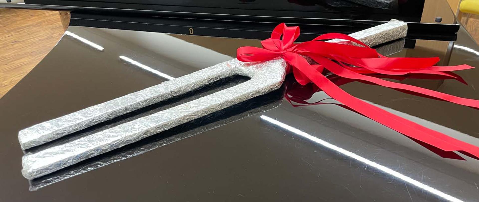 Na fortepianie leży przedmiot imitujący kamerton. Przedmiot jest pokryty folią aluminiową i przewiązany czerwoną wstążką.