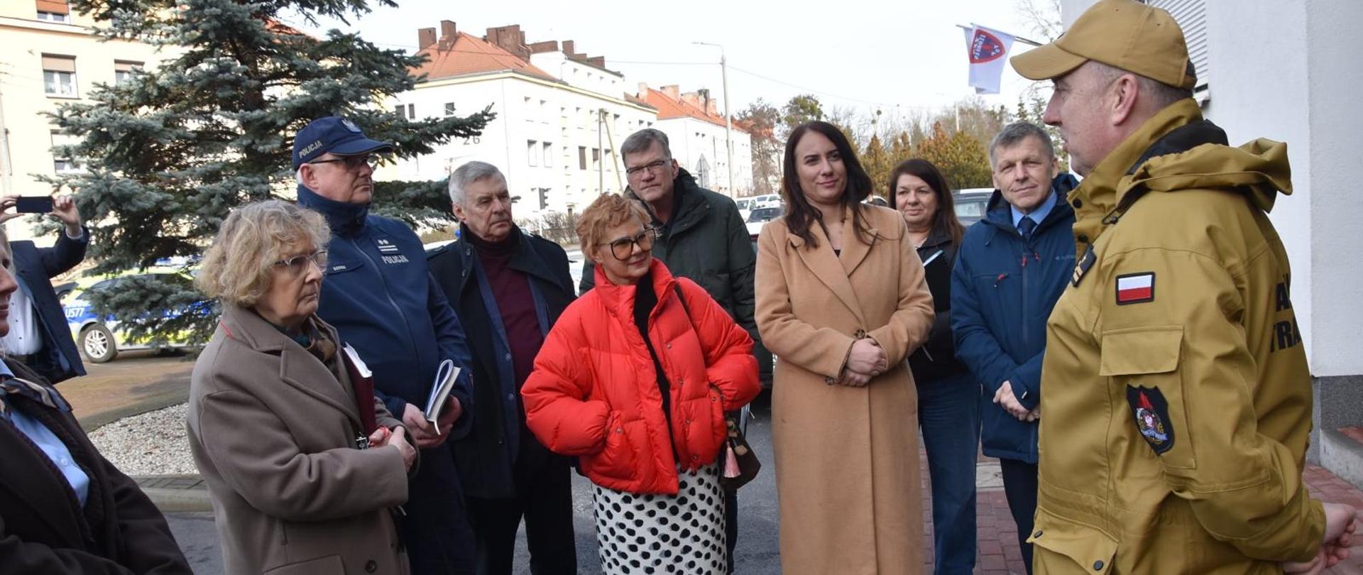 Na zdjęciu widać grupę kilkunastu osób stojących na zewnątrz, przed budynkiem, podczas spotkania lub krótkiej odprawy. Po prawej stronie stoi mężczyzna w piaskowym mundurze służbowym z naszywką Polski, który zwraca się do pozostałych uczestników. Naprzeciwko niego zgromadzona jest grupa kobiet i mężczyzn w płaszczach i kurtkach, ubranych stosownie do chłodnej pogody. Wśród nich widać także osoby w mundurach. W tle znajdują się drzewa, zaparkowane samochody oraz zabudowania mieszkalne. Atmosfera wygląda na oficjalną, a uczestnicy uważnie słuchają mówiącego.