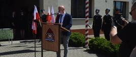 Plac przed komendą. Przy mównicy stoi przedstawiciel sekretarza stanu w MSWiA, ktory odczytuje zyczenia skierowane do strażaków przez ministra. W tle stojak z flagami oraz poczet flagowy.