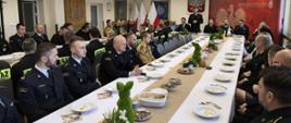 Przy stołach na których znajdują się dekoracje świąteczne i talerze, siedzą strażacy i zaproszenie goście. Strażacy ubrani są w mundury galowe zaproszeni goście odświętnie w garniturach. W tle logo Państwowej Straży Pożarnej oraz godło Polski i flagi