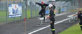 XI Powiatowe Zawody Sportowo-Pożarnicze OSP i MDP. Stadion w Miejskiej Górce. Na bieżni rozgrwana jest sztafeta pożarnicza 7x50 m z przeszkodami. Zawodnik pokonuje równoważnię. Ramiona ma rozłożone na boki. W prawej dłoni trzyma prądownicę. 
