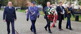 Wojewoda pomorski niesie wieniec obok idą zaproszeni goście oraz przedstawiciele Policji oraz Państwowej Straży Pożarnej.