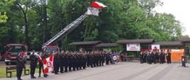 Strażacy stoją w szeregach w Parku im. Poległych Górników w Zabrzu. Za nimi znajdują się pojazdy pożarnicze, a z rozłożonej nad nimi drabiny mechanicznej powiewa flaga Polski. Na wprost nich gra Dęta Orkiestra Muzeum Górnictwa Węglowego w Zabrzu. W szeregach strażaków widoczne są poczty sztandarowe.