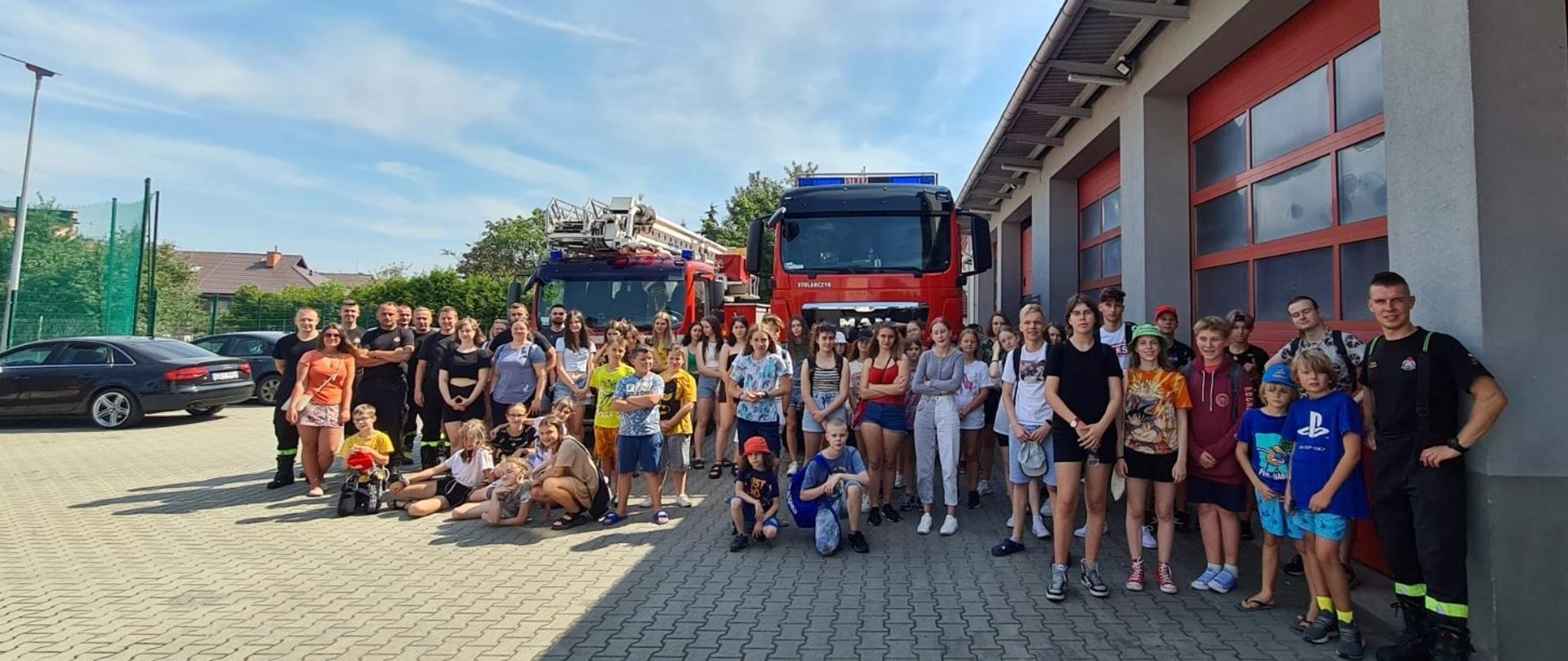 Pamiątkowe zdjęcie grupy dzieci z kolonii artystycznych z lęborskimi strażakami na placu Komendy Powiatowej Państwowej Straży Pożarnej w Lęborku. W tle dwa samochody :jeden ciężki ratowniczo - gaśniczy, drugi samochód specjalny podnośnik. 