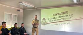 Na zdjęciu sala szkoleniowa. Na ścianie na ekranie rzutnika wyświetla się napis Szkolenie z zakresu działania rot asekuracyjnych "RIT". Na dole napis Sosnowiec 11-13 lipiec 2023 r. Po lewej stronie logo Państwowej Straży Pożarnej. Obok ekranu stoi strażak w piaskowym mundurze służbowym. Po lewej stronie, pod oknem siedzi czterech strażaków w czarnych mundurach koszarowych. Na pierwszym planie widoczny stoli na którym ustawiony jest rzutnik i laptop. Na stoliku leża papiery i stoi butelka wody mineralnej. Obok stolika stoi strażak w pisakowym mundurze służbowym.