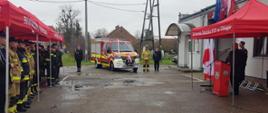 W centralnej części zdjęcia stoi nowy pojazd pożarniczy przyozdobiony biało-czerwonymi wstęgami. Obok auta i po lewej stronie, pod czerwonym namiotem stoją strażacy w mundurach. Po prawej stronie tuż przed remizą osp stoi czerwony namiot z mównicą, pod którym zgromadzenia są zaproszenia goście.