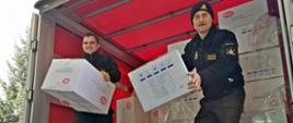 Zdjęcie z przodu. Dwaj strażacy PSP w trakcie rozładunku pudełek z maseczkami ochronnymi z pojazdu ciężarowego. 