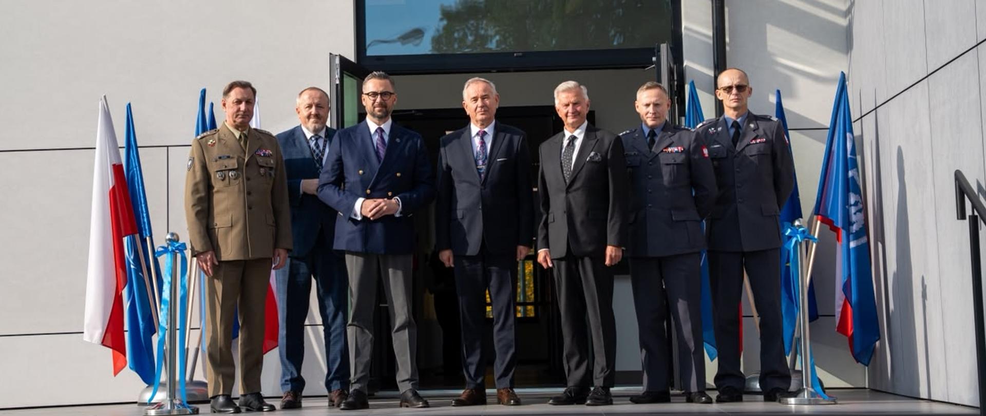 Na dworze, przed wejściem do budynku ze ścianami z szarych kafli stoi grupa siedmiu mężczyzn - trzech w mundurach, czterech w garniturach, trzeci od lewej minister Kulasek, za nimi po bokach flagi Polski i NATO w stojakach.
