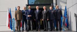 Na dworze, przed wejściem do budynku ze ścianami z szarych kafli stoi grupa siedmiu mężczyzn - trzech w mundurach, czterech w garniturach, trzeci od lewej minister Kulasek, za nimi po bokach flagi Polski i NATO w stojakach.